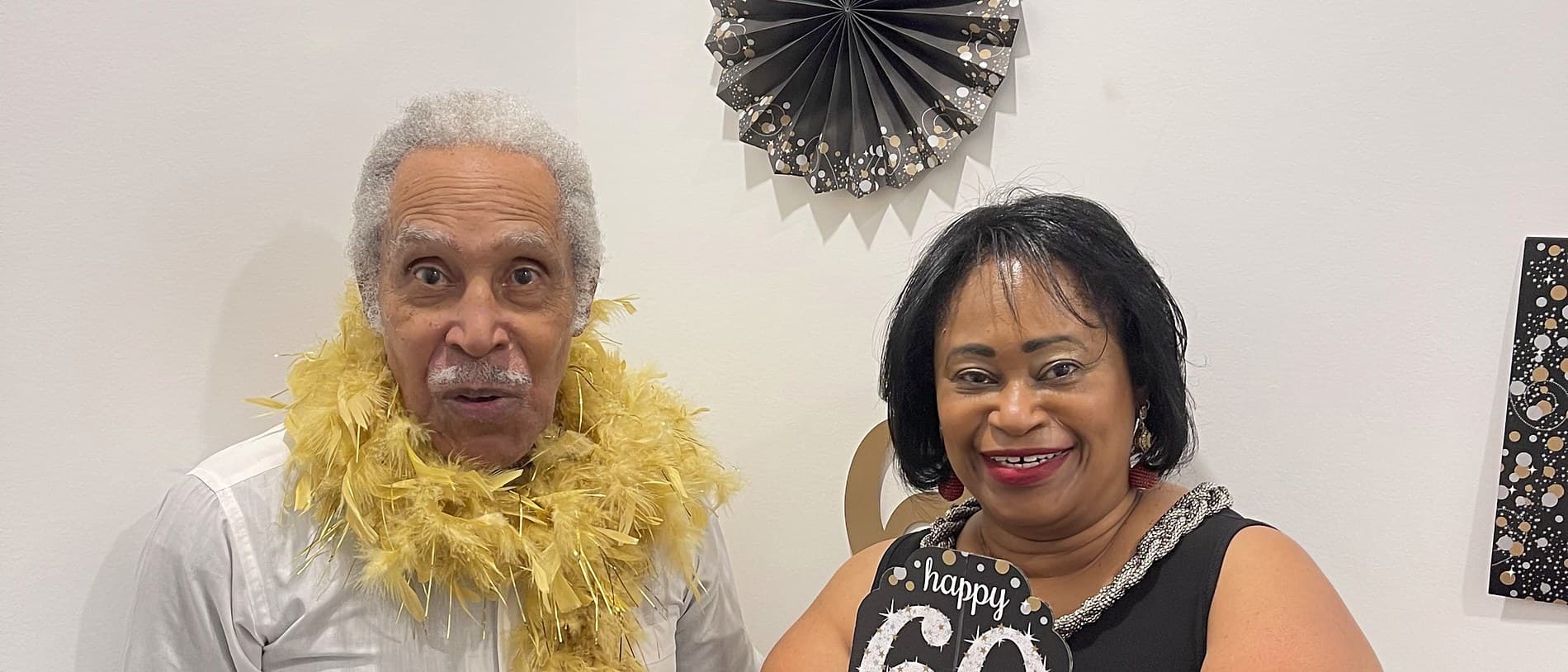 The owners together at a sixtieth birthday in the house, both holding party signs, one wearing a feather boa, paper fans on the wall behind them