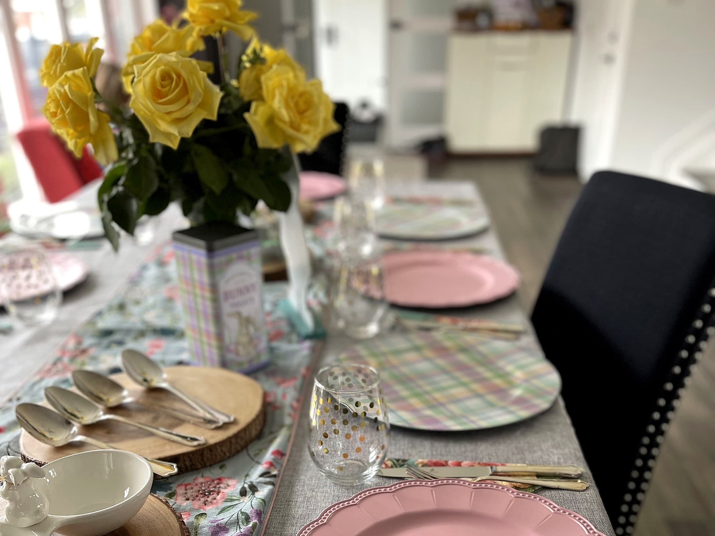 The dining table laid for an Easter gathering, with yellow roses, wood rounds and pastel place settings running the length of it