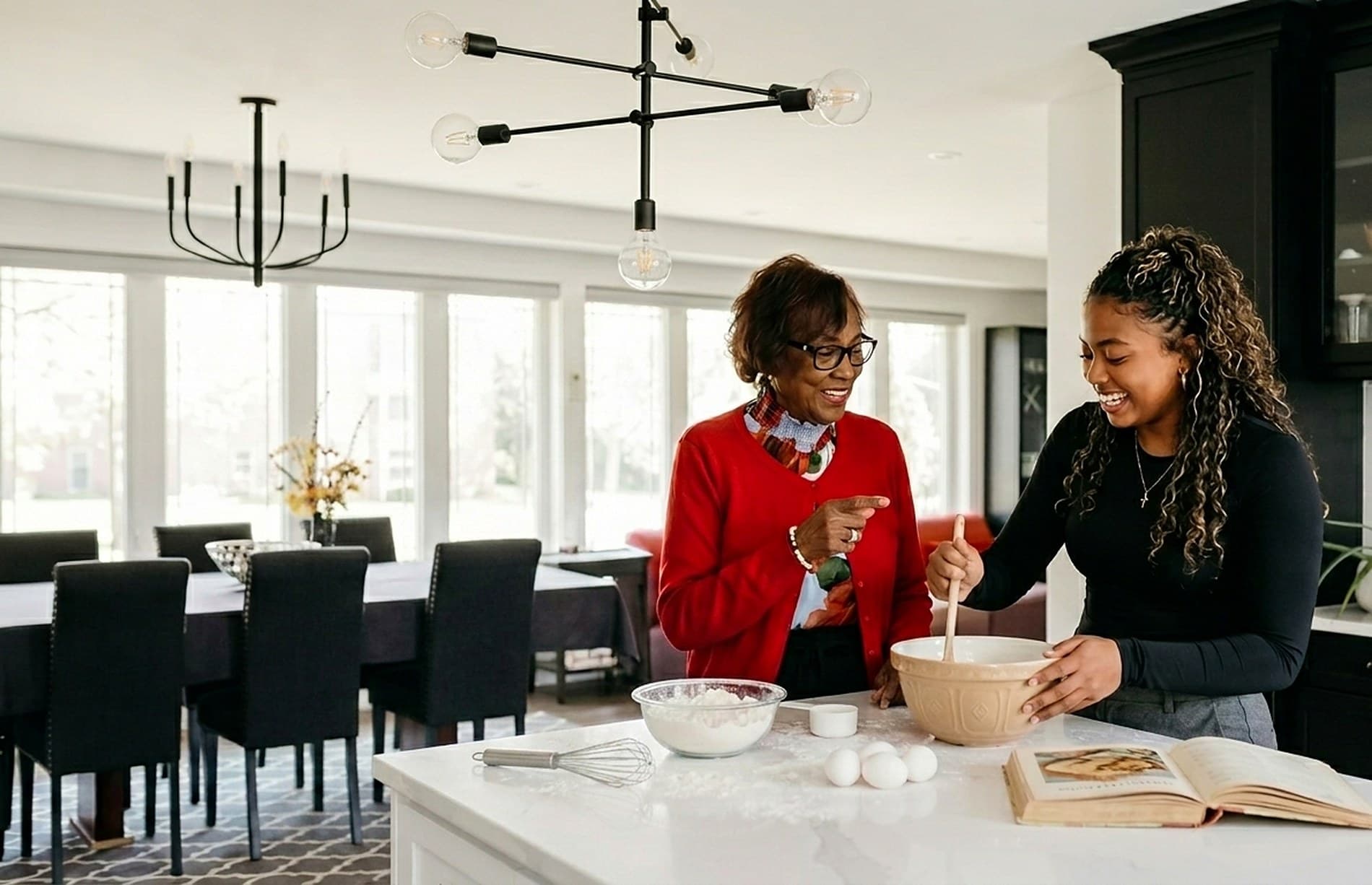 Two generations baking together at the kitchen island — a mixing bowl, a whisk, eggs and flour on the counter and an open cookbook beside them, with the long dining table and the windows behind
