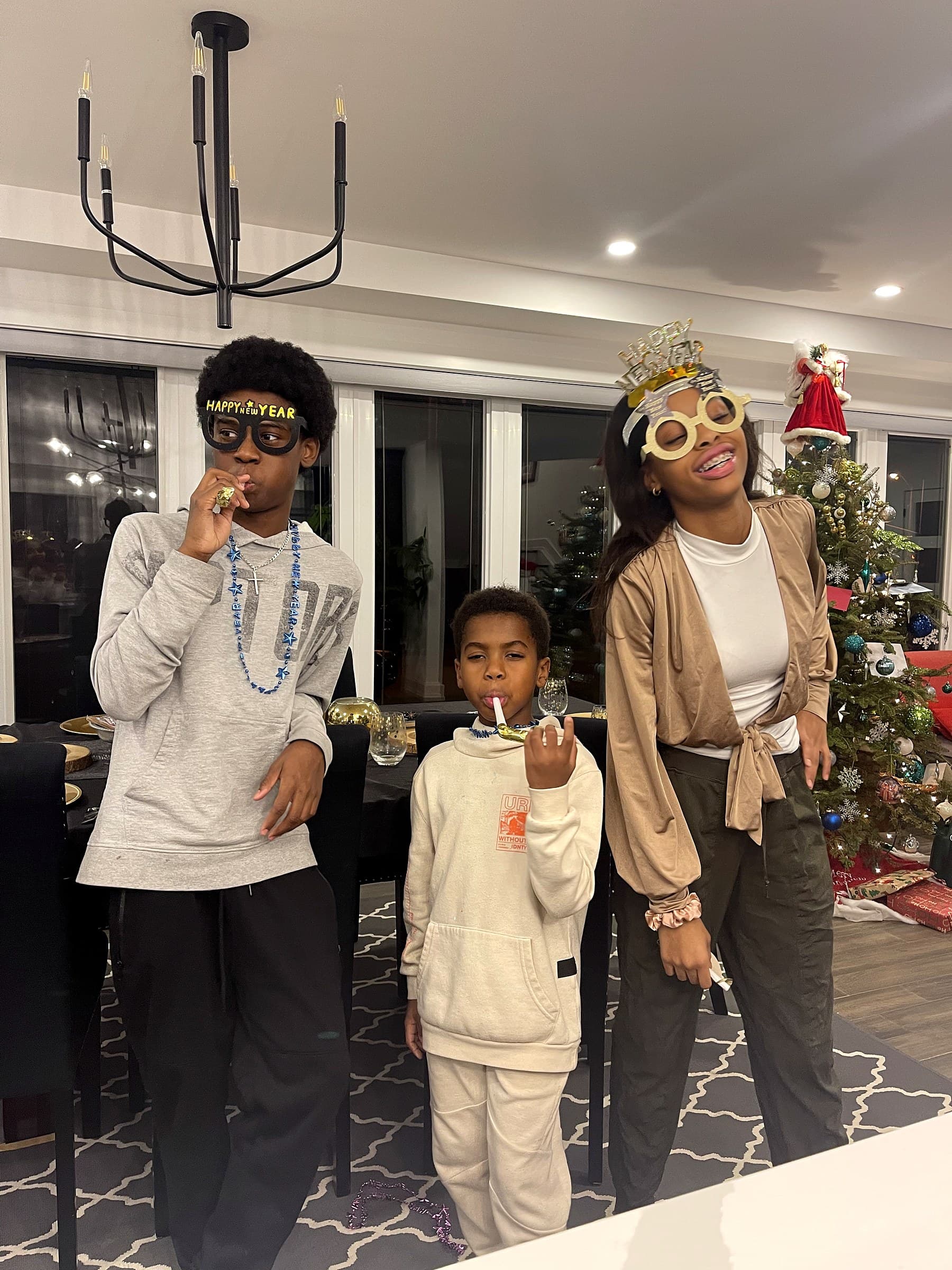 Three young people in Happy New Year glasses with party blowers, a lit Christmas tree behind them