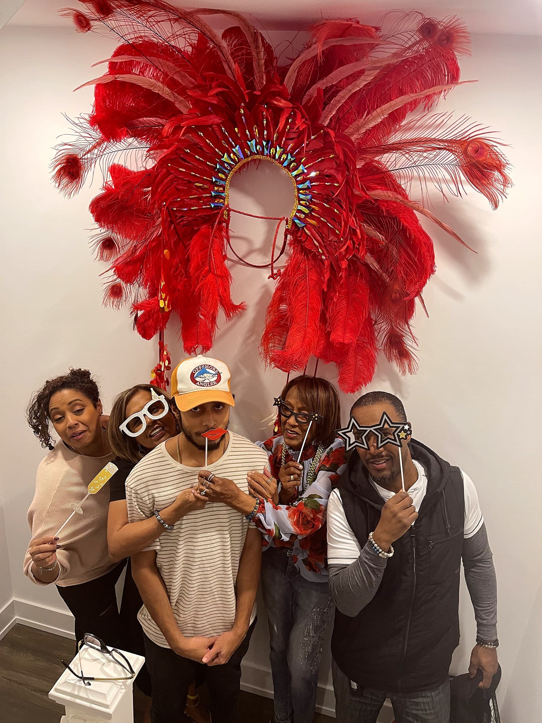 Five adults posing with photo-booth props beneath a large red carnival feather headdress mounted on the wall