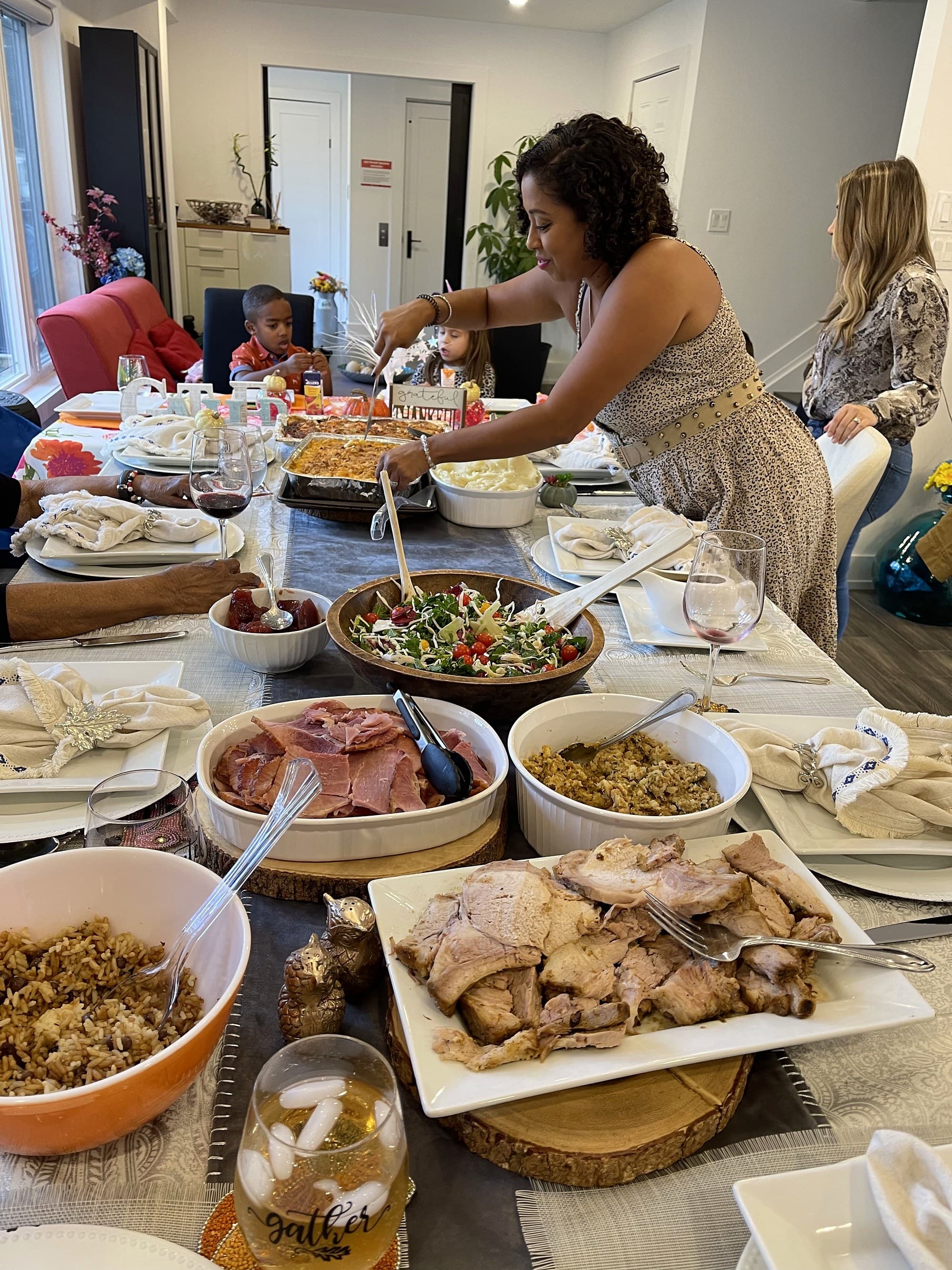 A family meal being served: a table laid end to end with a ham, a roast, rice, salad and wine, someone serving from the far side and more of the family seated around it