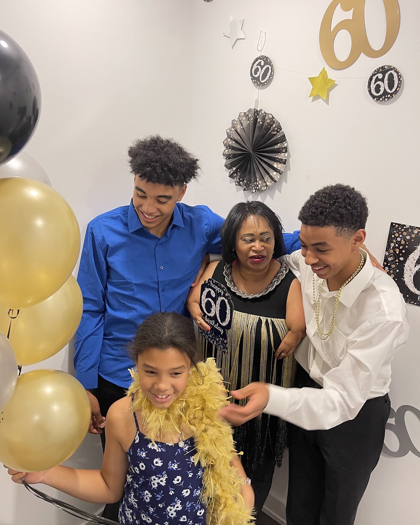 Three generations at a sixtieth birthday in the house: a grandmother in the centre with two grandsons either side and a granddaughter in front, gold and black balloons alongside them