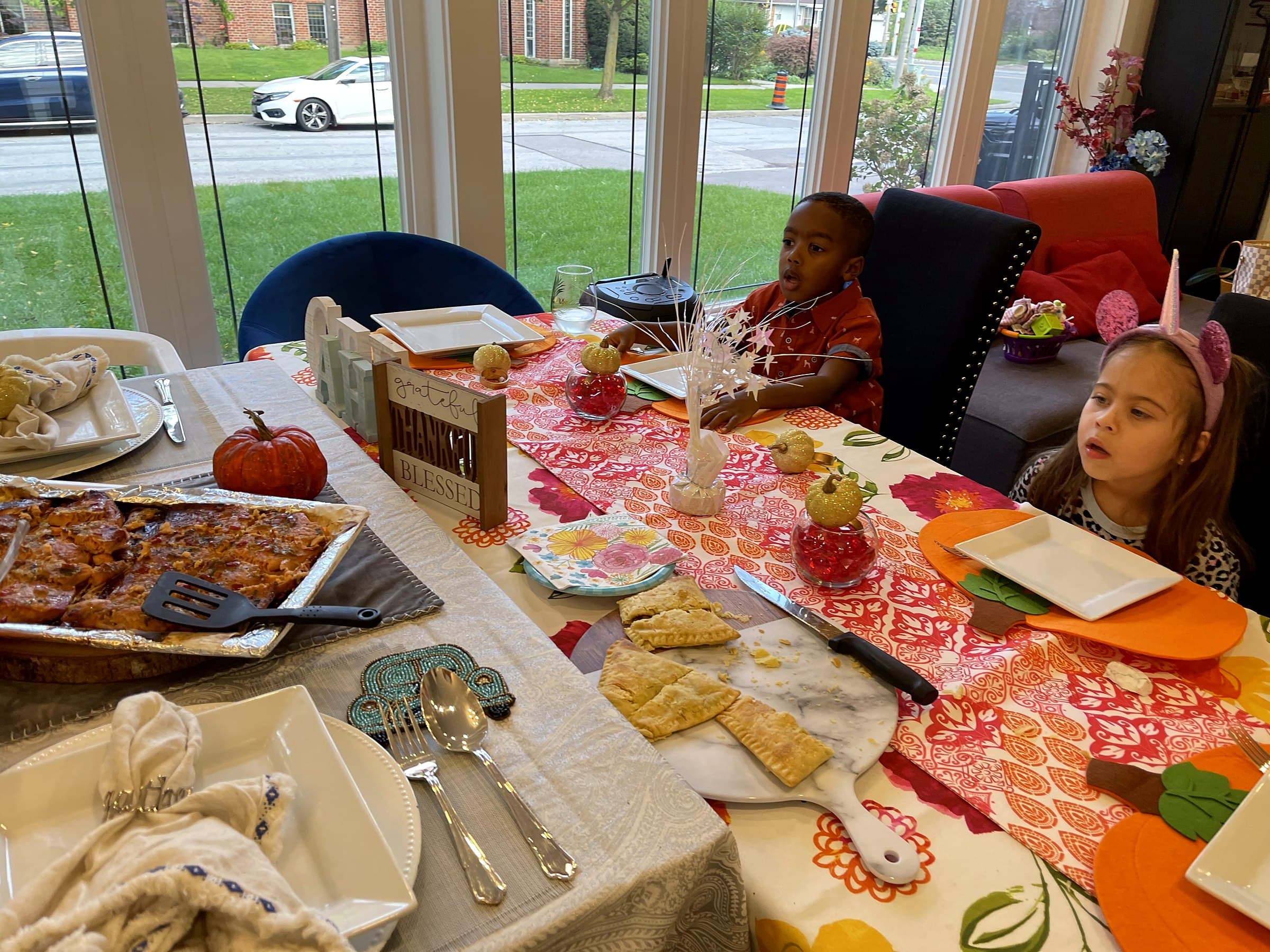 The long table set for Thanksgiving with an orange runner, gold pumpkins and a grateful, thankful, blessed sign, two children waiting at the far end