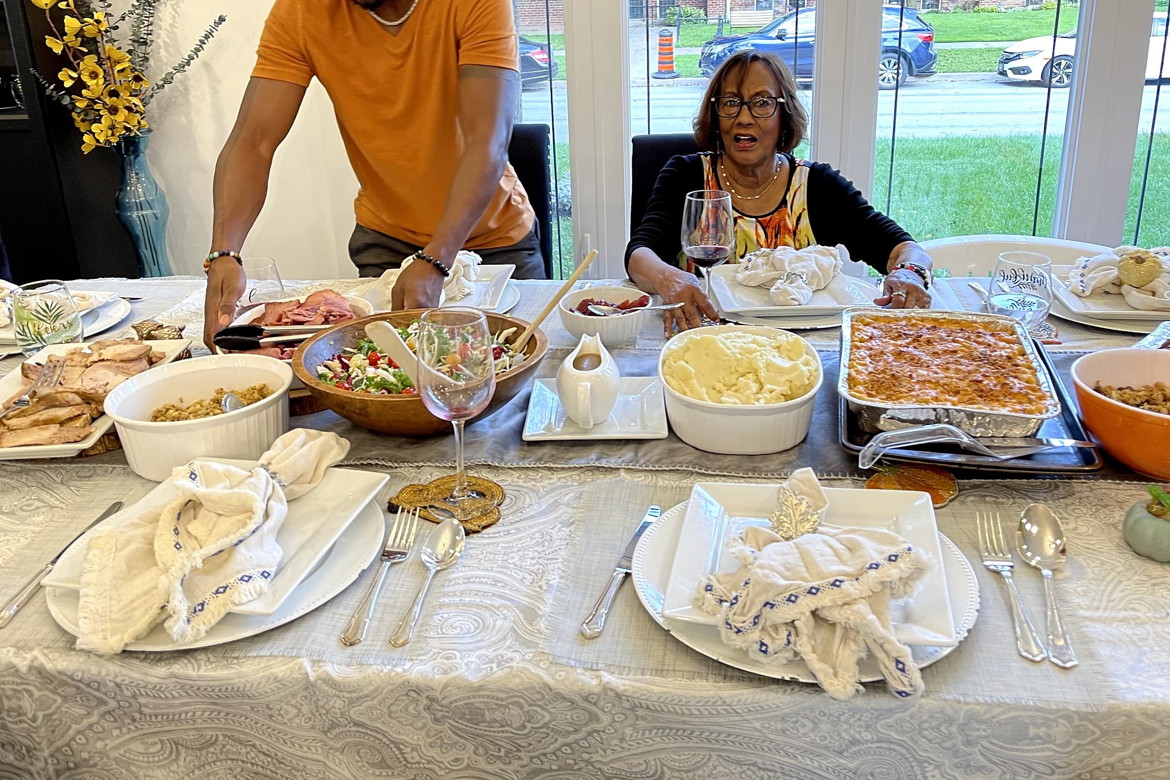 Three generations of a family at the Gatherwell dining table — a grandmother seated and smiling while a younger family member serves from a full spread of dishes
