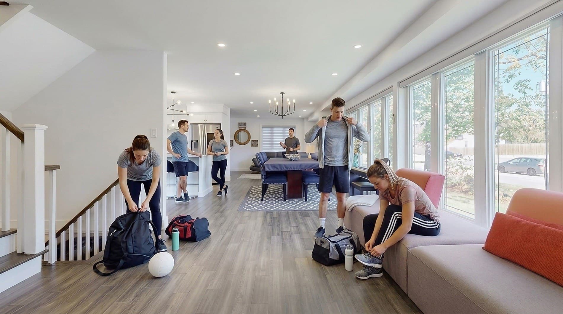 A team getting ready across the open main level: one player zipping a kit bag by the stairs beside a volleyball, another pulling on a jacket, a third lacing her shoes on the window bench, and more of the group talking by the kitchen island beyond